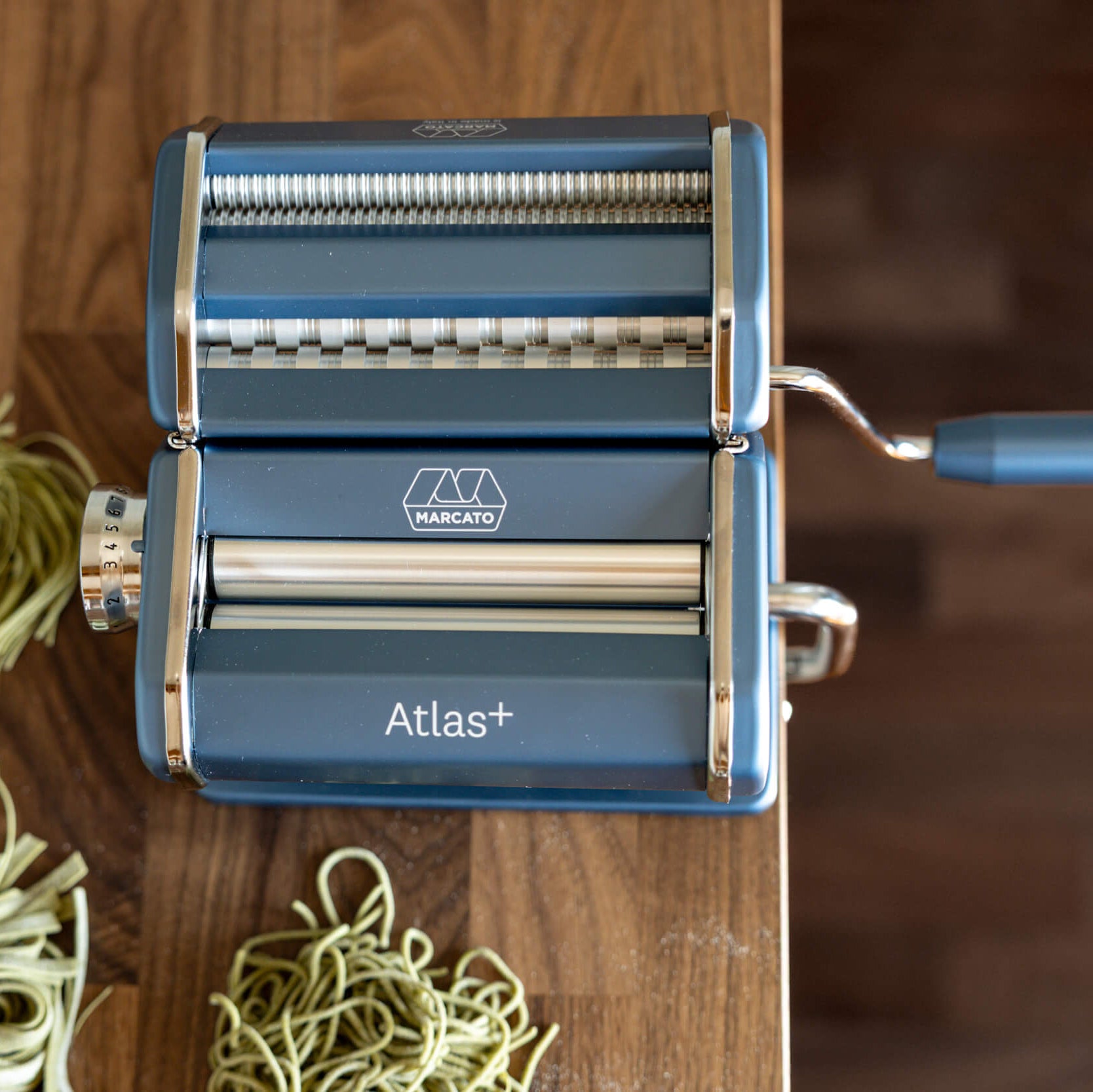 Blue pasta maker with 'Atlas+' branding on a wooden surface with fresh pasta strands.