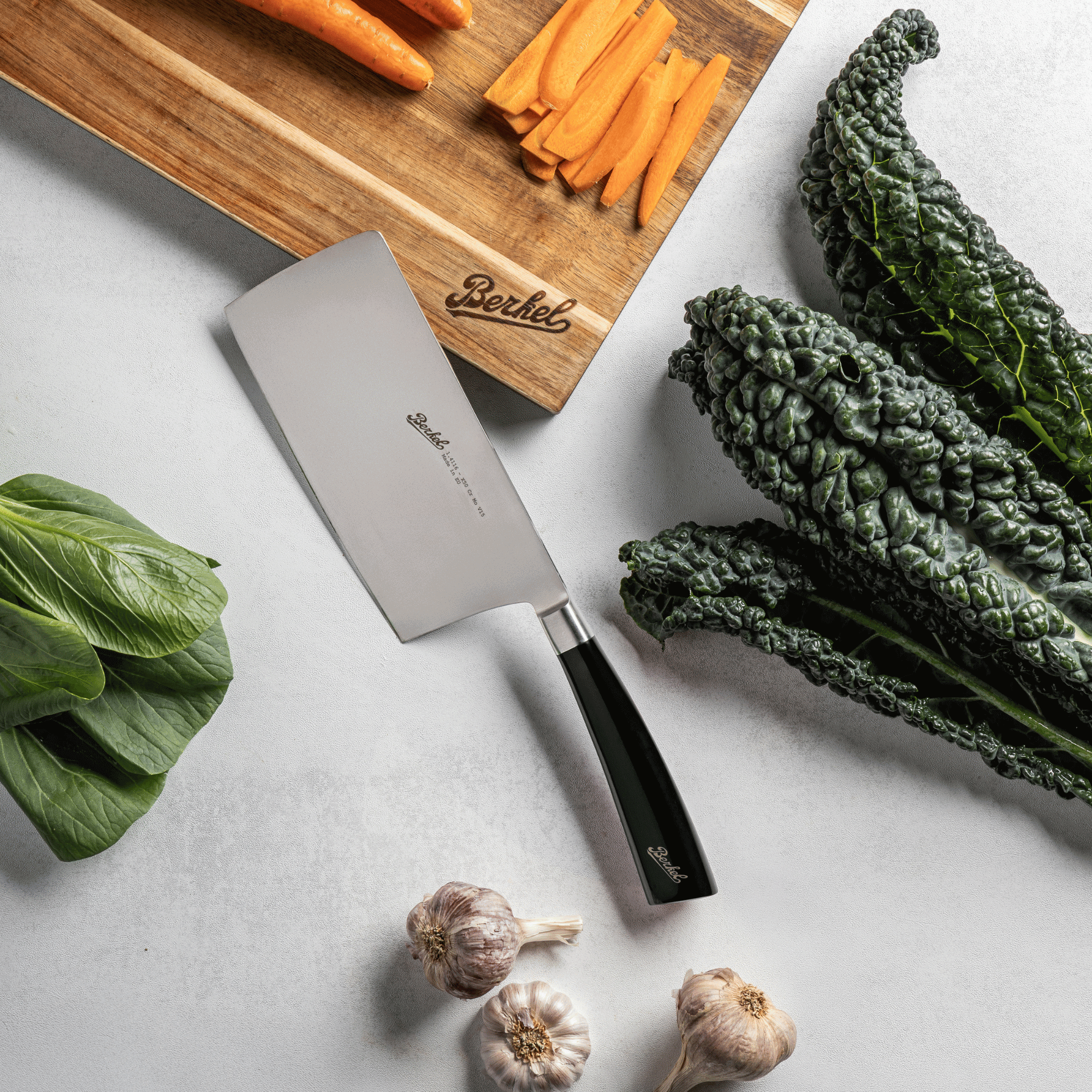 Vegetables on a cutting board with a Berkel Chinese chef's knife, including carrots, kale, and garlic, on a light background.