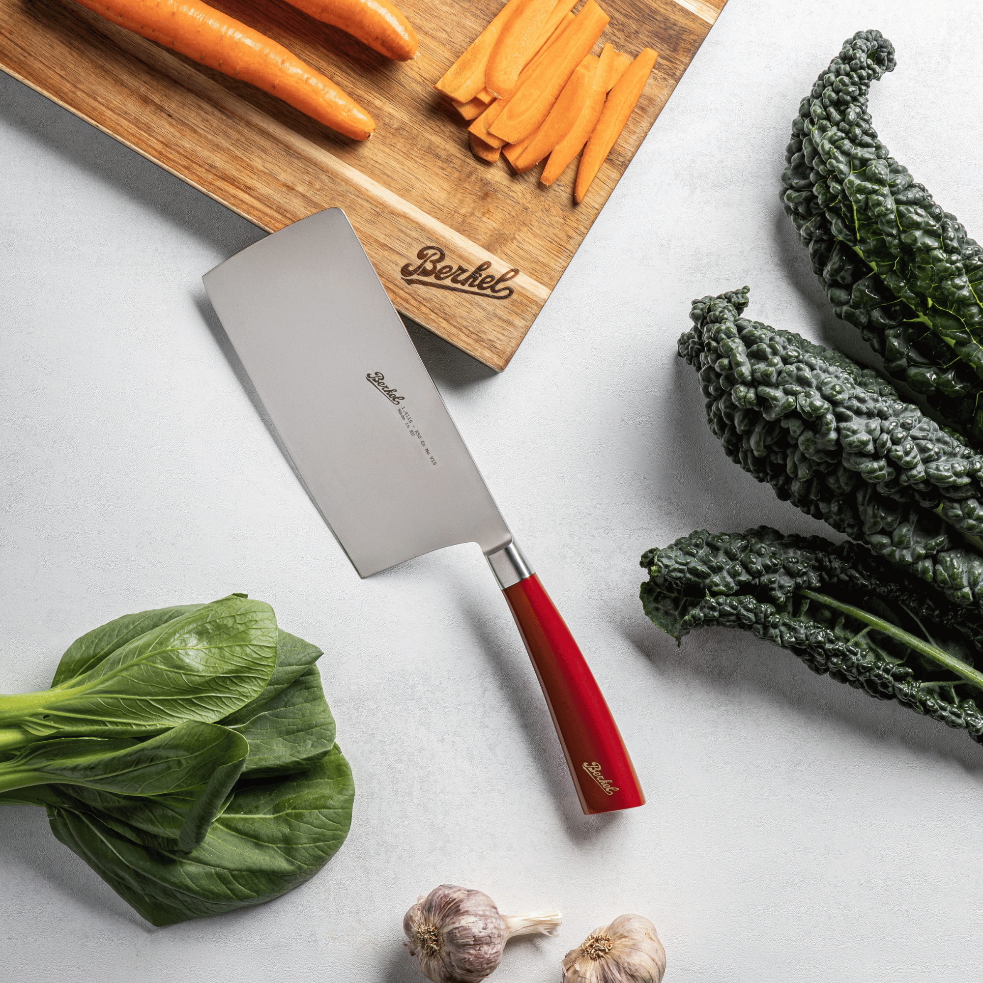 Vegetables on a cutting board with a Berkel Chinese chef's knife, including carrots, kale, and garlic, on a light background.