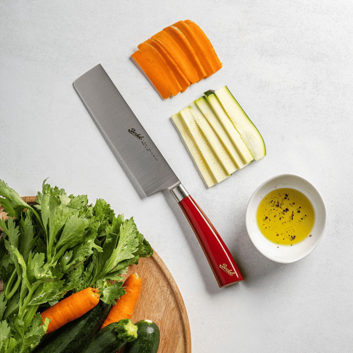 Red Berkel Elegance Nakiri knife next to cut cucumber, carrot and oil. 
