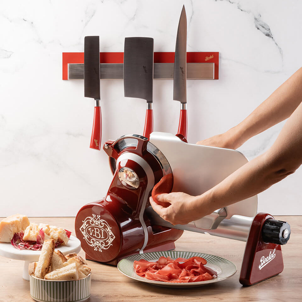 Berkel Icon 170 Slicer in Red in front of a marble wall and Berkel elegance knives. In use cutting cured meats.